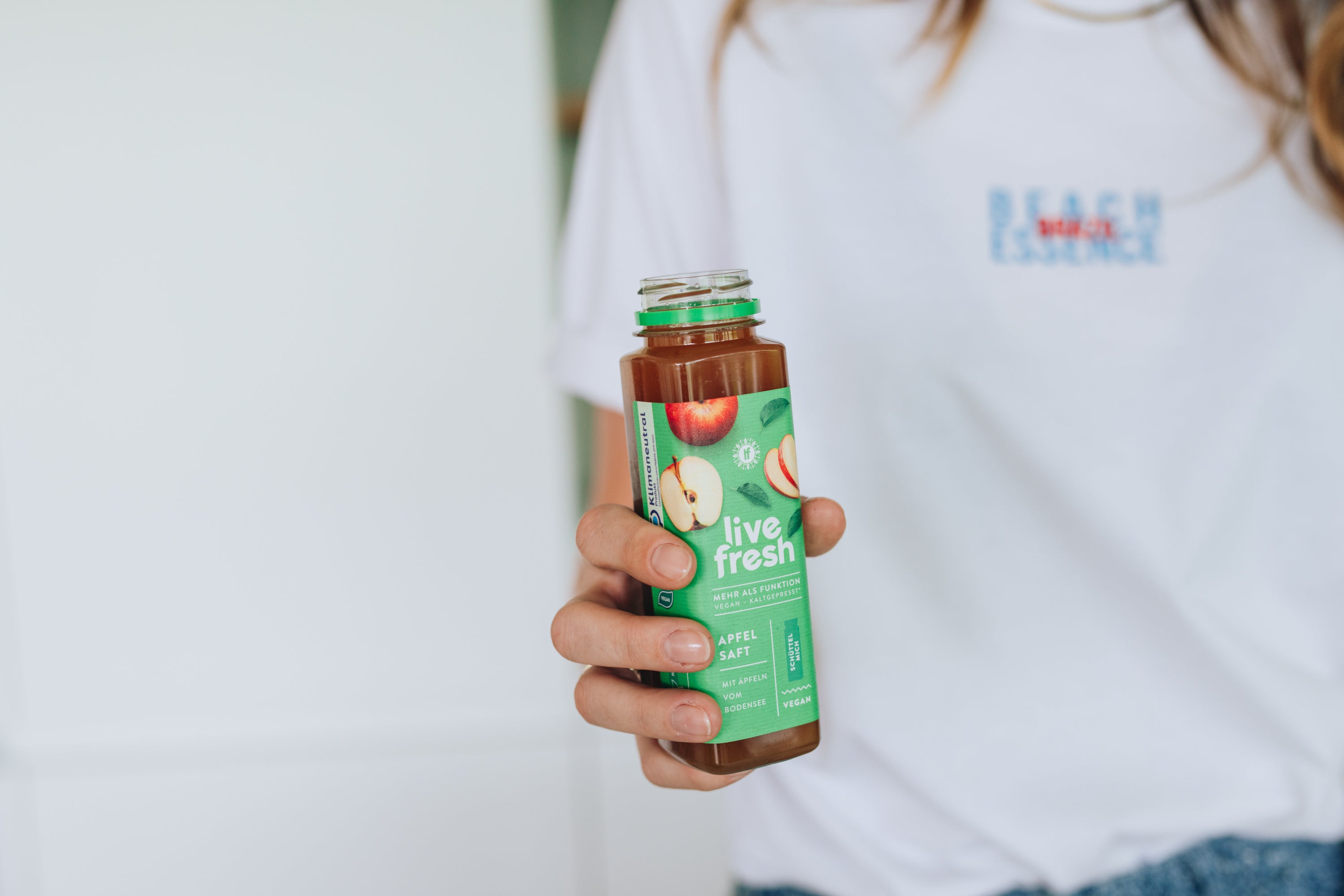 Person in weißem T-Shirt hält eine geöffnete Flasche „Live Fresh“ Apfelsaft mit grünem Etikett und Apfelgrafiken.