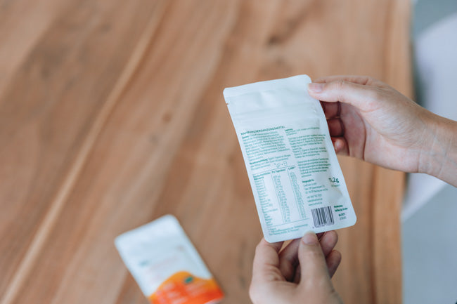 Nahaufnahme von zwei Händen, die eine Verpackung von Nahrungsergänzungsmitteln halten und die Rückseite lesen, auf der detaillierte Informationen zum Produkt, wie Inhaltsstoffe und Nährwertangaben, aufgedruckt sind. Auf dem Tisch im Hintergrund liegt eine weitere Verpackung eines ähnlichen Produkts, teils unscharf dargestellt. Der Tisch besteht aus Holz, was der Szene eine natürliche und entspannte Atmosphäre verleiht.