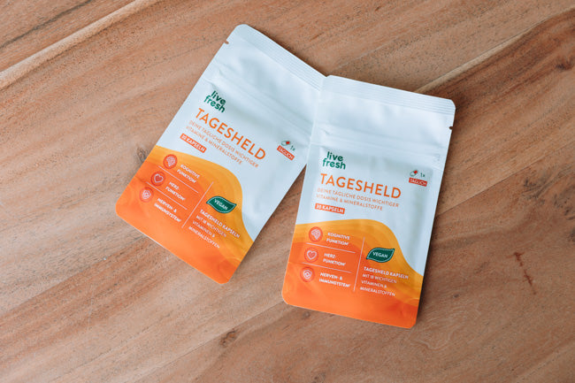 Zwei Packungen „Live Fresh Tagesheld“ Nahrungsergänzungskapseln, die auf einer Holzoberfläche liegen. Die Verpackungen sind in einem hellen Orange- und Weißton gehalten und enthalten Informationen über die Vorteile des Produkts, darunter „Kognitive Funktion“, „Immunsystem“, „Nervensystem“ und „Energie“. Die Kapseln sind als vegan gekennzeichnet und versprechen eine tägliche Versorgung mit wichtigen Nährstoffen.