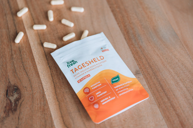 Packung „Live Fresh Tagesheld“ Nahrungsergänzungskapseln auf einer Holzoberfläche, umgeben von mehreren verstreuten Kapseln. Die Verpackung betont die Vorteile des Produkts, darunter die Unterstützung der „Kognitiven Funktion“, des „Nervensystems“ und des „Immunsystems“. Außerdem wird hervorgehoben, dass die Kapseln vegan sind. Die Szene vermittelt den Eindruck einer natürlichen und gesunden Ergänzung für den Alltag.