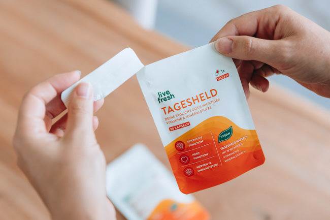 Zwei Hände, die gerade eine Packung „Live Fresh Tagesheld“ Nahrungsergänzungskapseln öffnen. Auf der Verpackung sind die Vorteile der Kapseln hervorgehoben, darunter Unterstützung für die „Kognitive Funktion“, das „Immunsystem“ und das „Nervensystem“. Zudem wird betont, dass die Kapseln vegan sind. Im Hintergrund ist eine weitere Packung des Produkts zu sehen. Die Szene zeigt den praktischen Gebrauch der Kapseln im Alltag.