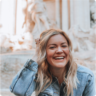 Blonde Frau im Jeanshemd, die lächelnd in die Kamera blickt und eine Hand in den Haaren hat. Im Hintergrund ist ein weißer Brunnen zu erkennen.