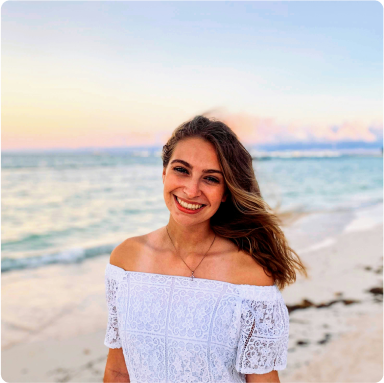 Lächelnde, brünette Frau im weißen Kleid. Im Hintergrund sind Strand und Meer zu sehen.
