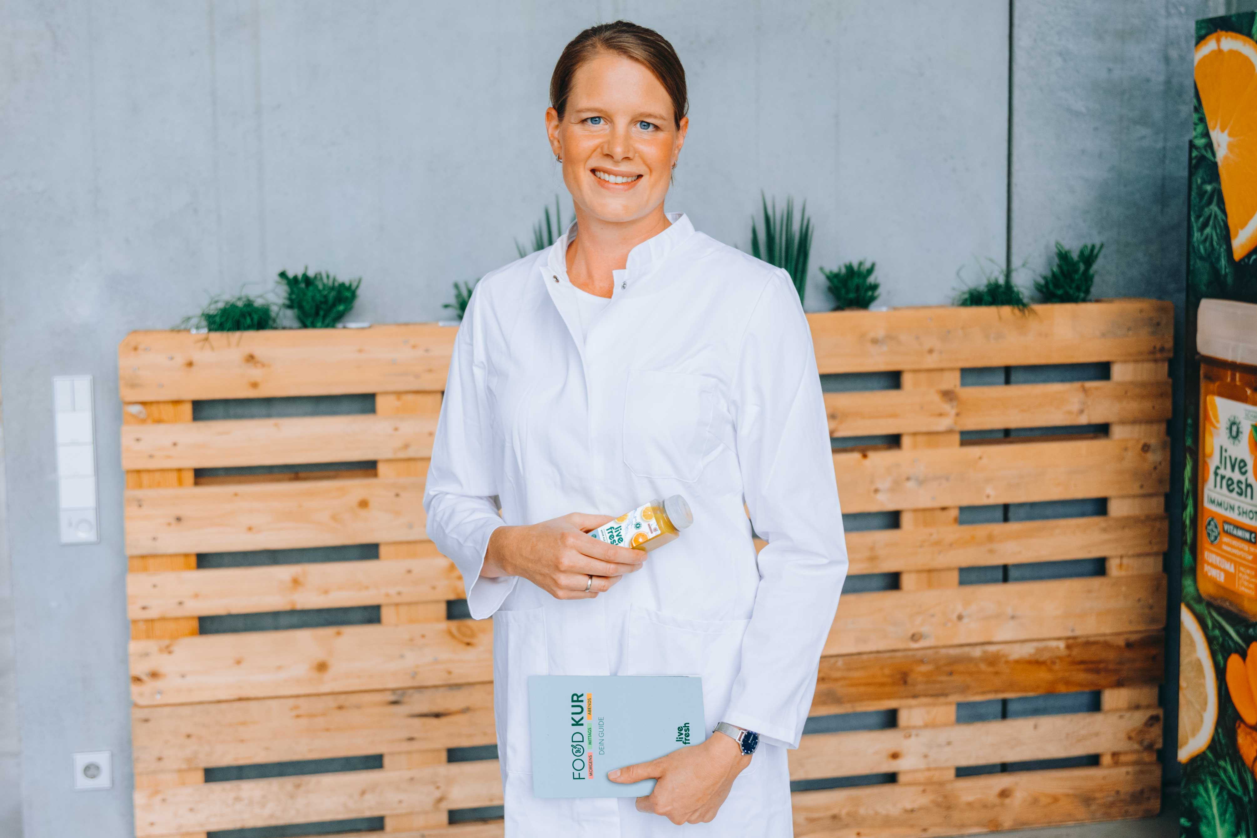 Eine lächelnde Frau mit braunen, zusammengebundenen Haaren in einem weißen Kittel hält eine Flasche Saft von LiveFresh in der einen Hand und ein Buch mit der Aufschrift