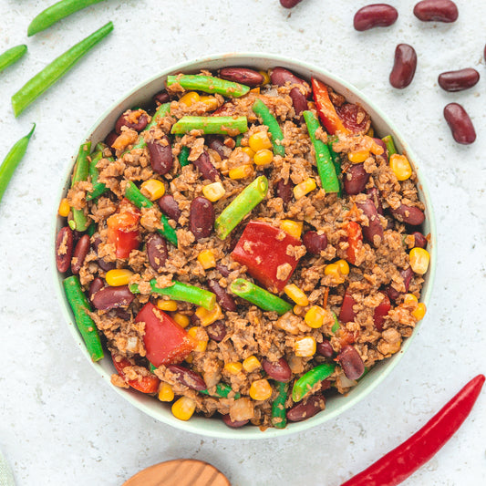Eine Schale mit "Chili sin Carne" von Live Fresh. Die vegane Mahlzeit enthält grüne Bohnen, Kidneybohnen, Mais, Tomaten und pflanzliches Hack. Um die Schale herum liegen frische Chilischoten und Bohnen, was die Zutaten des Gerichts hervorhebt.