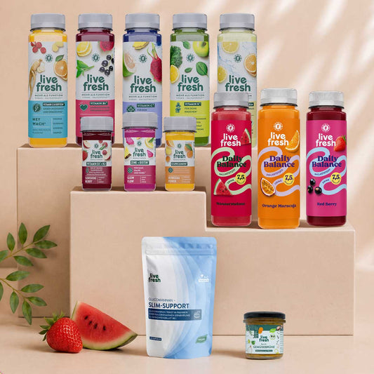 Mehrere Produkte der LiveFresh Saftkur sind auf beigefarbenen Podesten arrangiert. Im Mittelpunkt stehen drei Flaschen der Daily Balance Special Edition in den Sorten Wassermelone, Orange Maracuja und Red Berry, umgeben von weiteren Saftkur Flaschen, Nahrungsergänzungen und frischen Früchten wie Wassermelone und Erdbeere.