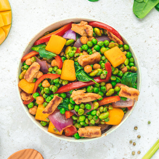 Eine bunte Bowl mit frischen Zutaten wie grünen Erbsen, roten Paprikastreifen, roten Zwiebeln, Kichererbsen, Spinat und goldgelben Mangostücken, ergänzt durch knusprige vegane Proteinstücke. Die Mischung vermittelt ein gesundes, ausgewogenes und farbenfrohes Gericht.