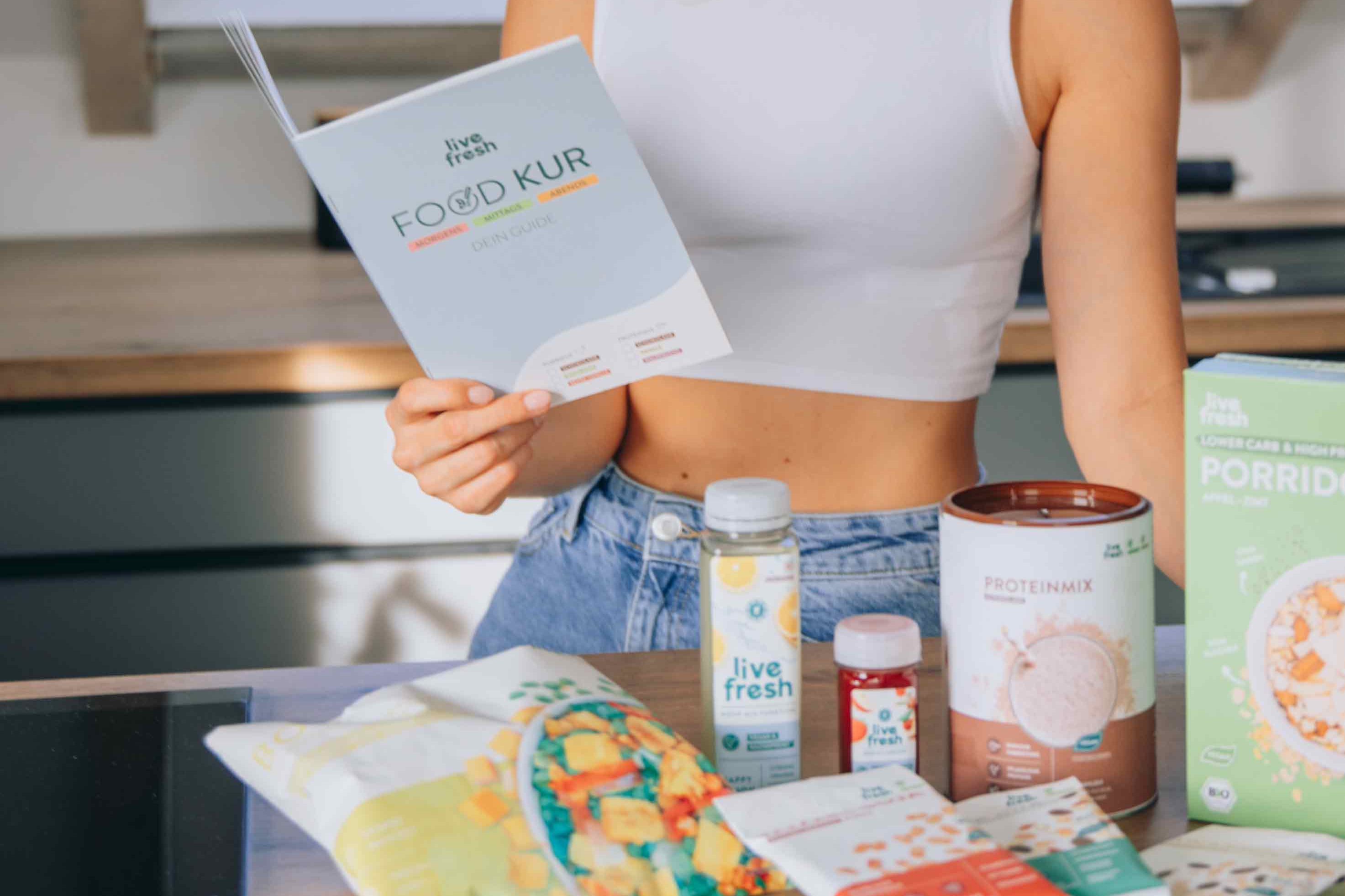 Eine Frau die in einem hellen, modernen Raum, die ein Heft mit der Aufschrift „Food Kur - Dein Guide“ von Live Fresh liest. Vor ihr auf dem Tisch liegen verschiedene Produkte von Live Fresh, darunter ein Proteinmix, ein Porridge-Pack, ein Saft, ein Nahrungsergänzungsmittel und eine Fertigmahlzeit. Sie trägt ein weißes, ärmelloses Oberteil und scheint sich auf die gesunde Ernährung im Rahmen des Food-Kur-Programms zu konzentrieren.