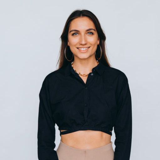 Junge Frau mit offenen, braunen Haaren, goldenen Creolen und Kette trägt ein schwarzes Cropped-Hemd und beige Leggings, während sie lächelnd in die Kamera schaut.