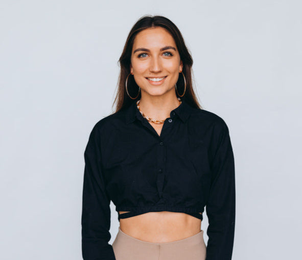 Junge Frau mit offenen, braunen Haaren, goldenen Creolen und Kette trägt ein schwarzes Cropped-Hemd und beige Leggings, während sie lächelnd in die Kamera schaut.