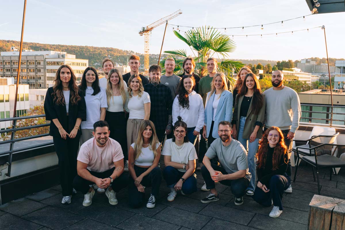 Gruppe von Menschen auf einer Dachterrasse bei Sonnenuntergang, mit Blick auf eine Stadtlandschaft mit Gebäuden, einem Kran und einer Palme.  Einige Personen hocken sich hin, die anderen stehen. Alle lächeln und posieren für ein gemeinsames Foto.
