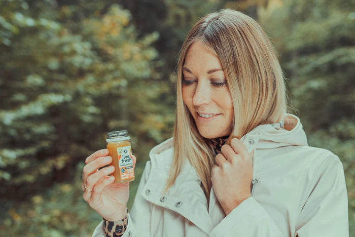 unge Frau mit blonder Frisur und heller Jacke steht im Grünen und hält einen kleinen LiveFresh-Shot in der Hand, während sie lächelnd darauf blickt.