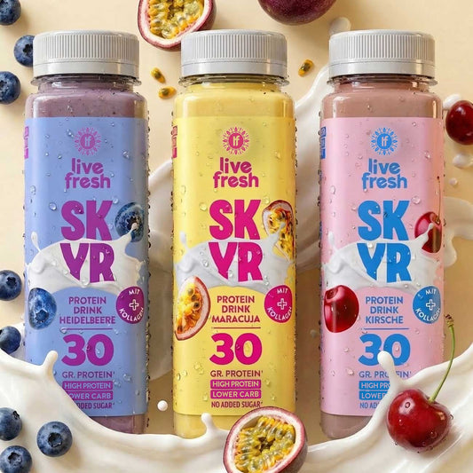 Drei Flaschen Skyr Protein Drink mit Blaubeere, Maracuja und Kirsche stehen vor einem cremigen Milchsplash, umgeben von den passenden Früchten. Auf den Etiketten steht „live fresh“, „SKYR“, „PROTEIN DRINK HEIDELBEERE“, „PROTEIN DRINK MARACUJA“, „PROTEIN DRINK KIRSCHE“, „MIT KOLLAGEN“, „30 GR. PROTEIN“, „HIGH PROTEIN“, „LOWER CARB“ und „NO ADDED SUGAR“.