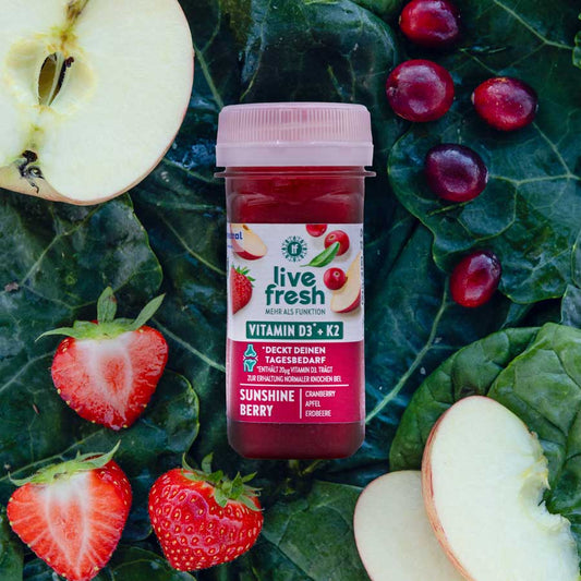 Sunshine Berry Shot von LiveFresh mit Viramin D3 + K2 Aufschrift. Obst und Gemüse im Hintergrund. Enthält Cranberry, Apfel und Erdbeere.