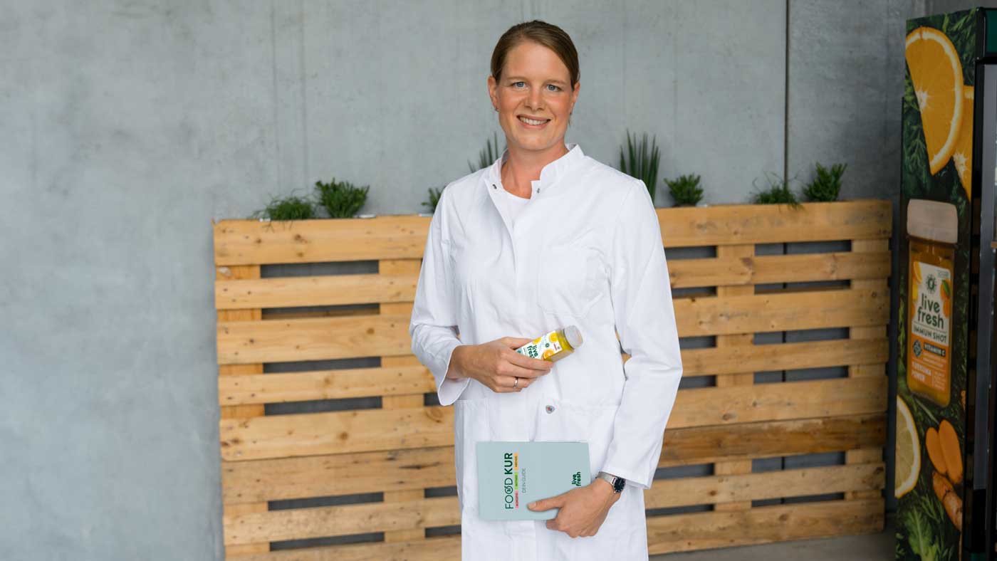 Eine lächelnde Frau mit braunen, zusammengebundenen Haaren in einem weißen Kittel hält eine Flasche Saft von LiveFresh in der einen Hand und ein Buch mit der Aufschrift "FOOD KUR" in der anderen Hand. Im Hintergrund stehen Pflanzen auf Holzpaletten vor einer grauen Wand.