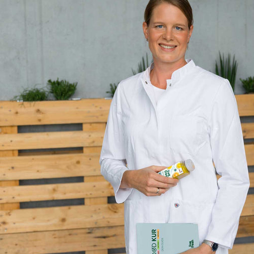 Eine lächelnde Frau mit braunen, zusammengebundenen Haaren in einem weißen Kittel hält eine Flasche Saft von LiveFresh in der einen Hand und ein Buch mit der Aufschrift