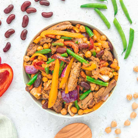 Bowl "Fajita Mexican Style" von LiveFresh, serviert in einer weißen Schüssel. Das Gericht enthält pflanzliches Geschnetzeltes, Kidneybohnen, Kichererbsen, Paprika, rote Zwiebeln und grüne Bohnen. Drumherum liegen frische Zutaten wie eine aufgeschnittene Paprika, grüne Bohnen, Kidneybohnen und Kichererbsen auf einer hellen Oberfläche. Ein Holzlöffel und ein Küchentuch liegen am unteren linken Rand.