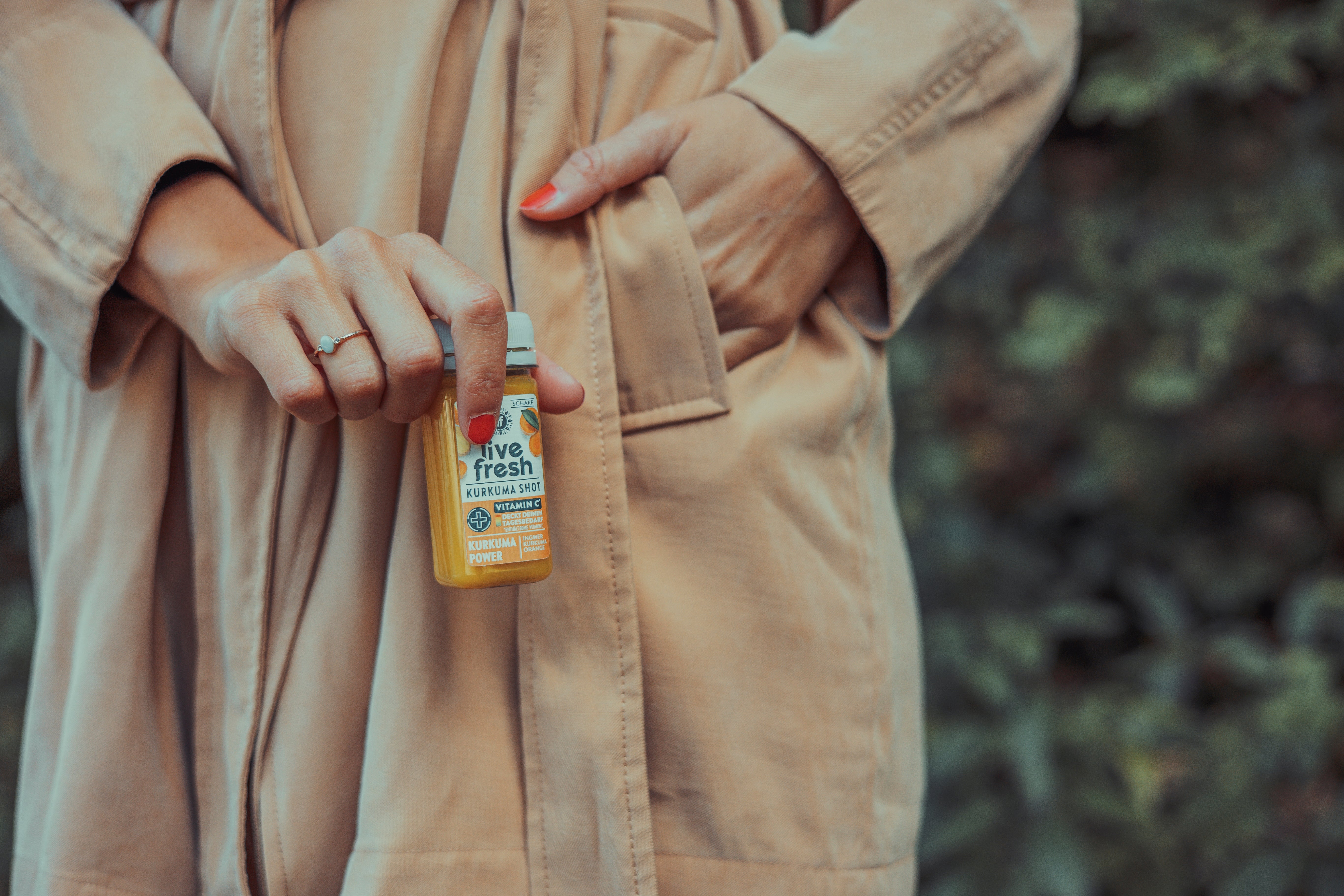 Nahaufnahme einer Frauenhand, die eine kleine Flasche Live Fresh Kurkuma Shot hält, wobei das Etikett vor dem Hintergrund ihres beigen Trenchcoats und einer natürlichen, begrünten Umgebung gezeigt wird.