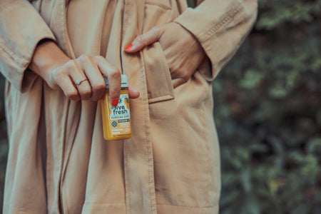 Nahaufnahme einer Frauenhand, die eine kleine Flasche Live Fresh Kurkuma Shot hält, wobei das Etikett vor dem Hintergrund ihres beigen Trenchcoats und einer natürlichen, begrünten Umgebung gezeigt wird.