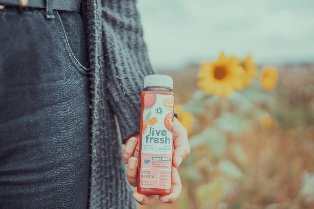 Hand hält eine Flasche rote Beete Saft vor einem Feld mit blühenden Sonnenblumen.