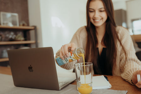 Eine Frau sitzt lächelnd vor einem Laptop und gießt frisch gepressten Orangensaft aus einer LiveFresh-Flasche in ein Glas.