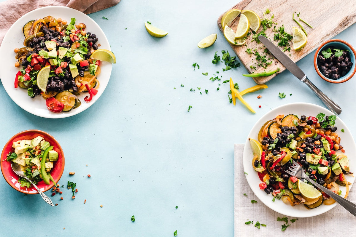 Ein gedeckter Tisch mit bunten, gesunden Gerichten zum Fastenbrechen, darunter Salate mit schwarzen Bohnen, Avocado, und Limetten auf weißem Geschirr und hellem Hintergrund.