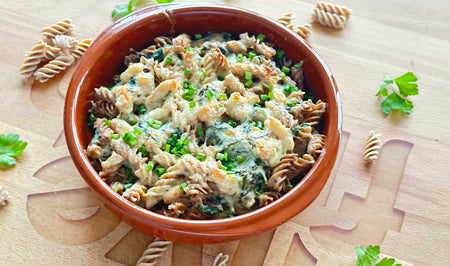 Überbackener Low Carb High Protein Nudelauflauf mit Vollkornfusilli, Spinat und cremiger Käsesauce, garniert mit frisch gehackten Kräutern, serviert in einer terrakottafarbenen Schale auf einem Holzbrett.