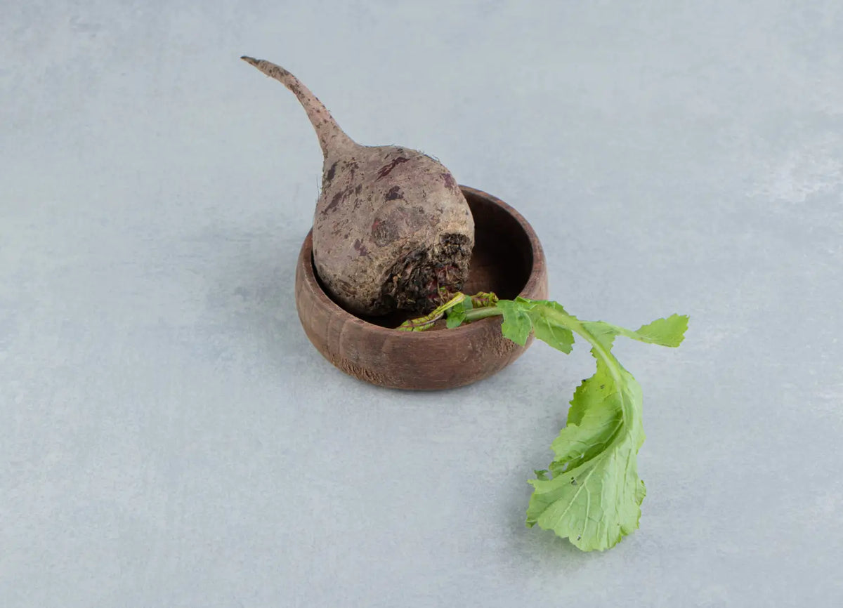 Rohe Maca-Wurzel mit grünen Blättern in einer kleinen Holzschale auf einer hellgrauen Oberfläche.