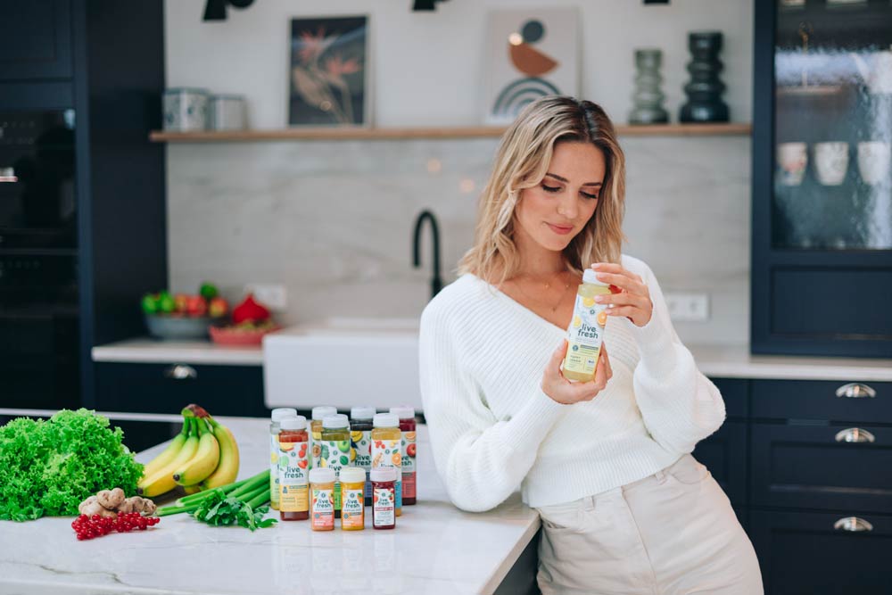 Eine Frau in einer modernen Küche, die eine Flasche „Live Fresh“-Saft in der Hand hält und das Etikett betrachtet. Vor ihr auf der Arbeitsplatte stehen mehrere weitere Flaschen des Saftes in verschiedenen Sorten sowie frische Zutaten wie Salat, Bananen, Ingwer und Frühlingszwiebeln. 