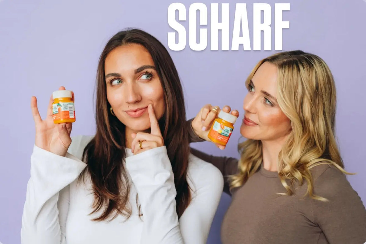 Zwei Frauen präsentieren orangefarbene Liv Fresh Ingwershots vor lilafarbenem Hintergrund. Die brünette Frau legt nachdenklich einen Finger an ihre Wange, während die blonde Frau sie anlächelt. Über ihnen steht "SCHARF".