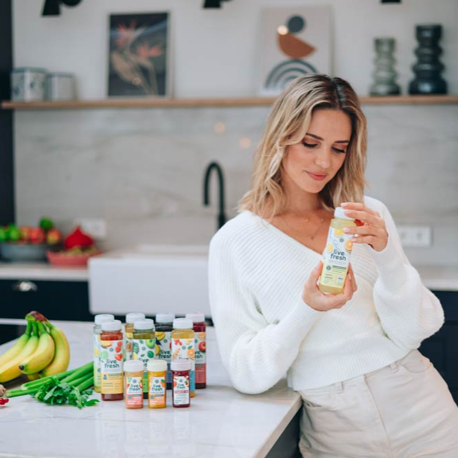 Eine junge Frau steht in der Küche mit Säften von LiveFresh neben sich und einem Happy Lemon Saft in der Hand. 