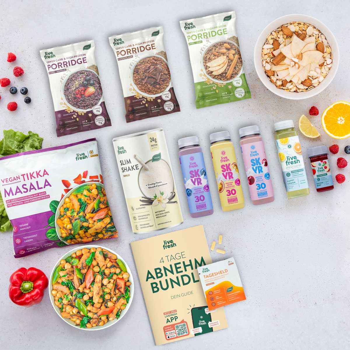 Flatlay mit dem „4 Tage Abnehm Bundle“ von LiveFresh, bestehend aus veganem Tikka Masala, Vanille Slim Shake, drei Skyr Protein Drinks (Heidelbeere, Maracuja, Kirsche), zwei Porridge-Sorten (Apfel-Zimt & Schoko), einem grünen Saft „Happy Lemon“, einem Vitamin D Shot sowie dem „Tagesheld“-Supplement. Daneben ein gedruckter Guide, frisches Obst und eine Bowl mit Nüssen und Apfelscheiben.
