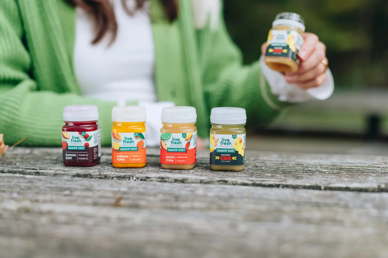 Vier LiveFresh Immunshots stehen auf einem rustikalen Holztisch im Freien. Im Hintergrund hält eine Frau mit grüner Strickjacke einen geöffneten Ingwer Xtreme Shot in der Hand. Die weiteren Sorten sind Sunshine Berry. Kurkuma Power und Ingwer Sturm.