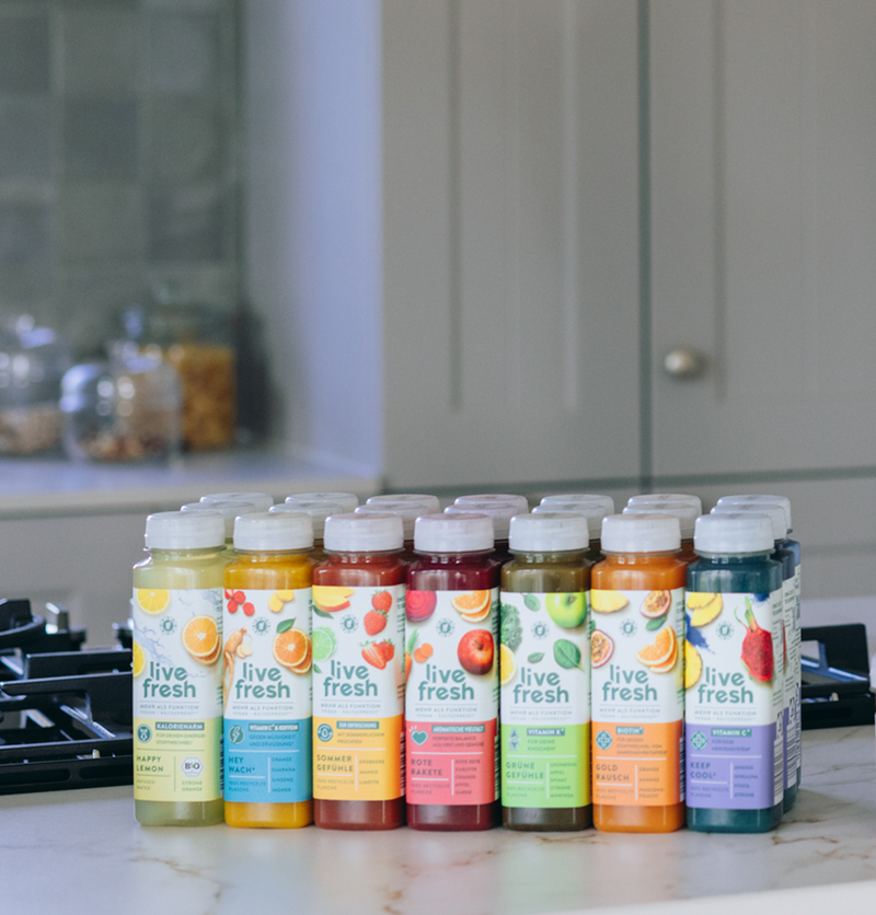 Mehrere bunte LiveFresh-Saftflaschen und Shots in verschiedenen Sorten (Happy Lemon, Hey Wach, Sommer Gefühle, Rote Rakete, Grüne Gefühle, Gold Rausch, Keep Cool) stehen aufgereiht auf einer hellen Küchenarbeitsplatte neben einem Herd. Im Hintergrund ein modernes Küchen-Setting.