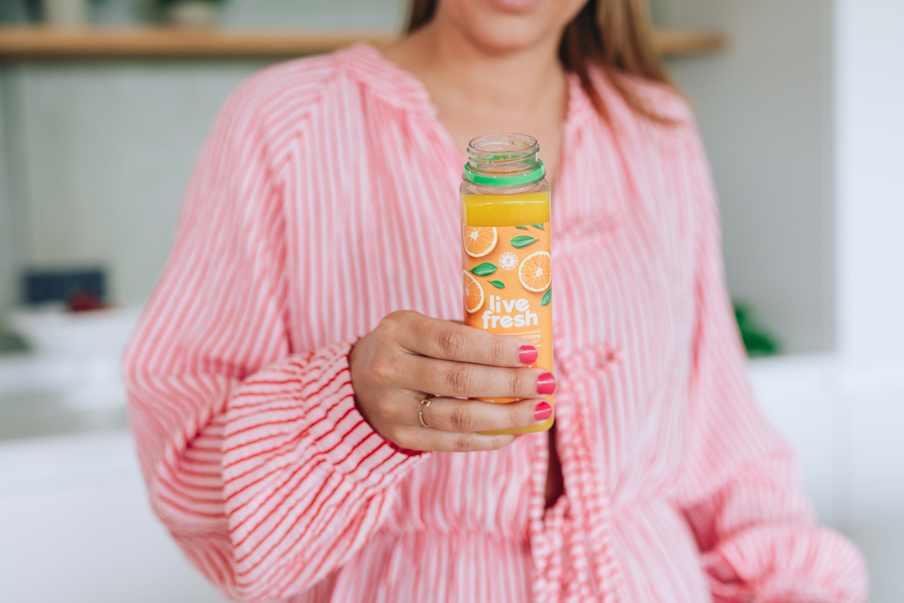 Frau in rosa-weiß gestreifter Bluse hält eine geöffnete Flasche „Live Fresh“ Orangensaft mit Orangenmotiv auf dem Etikett.