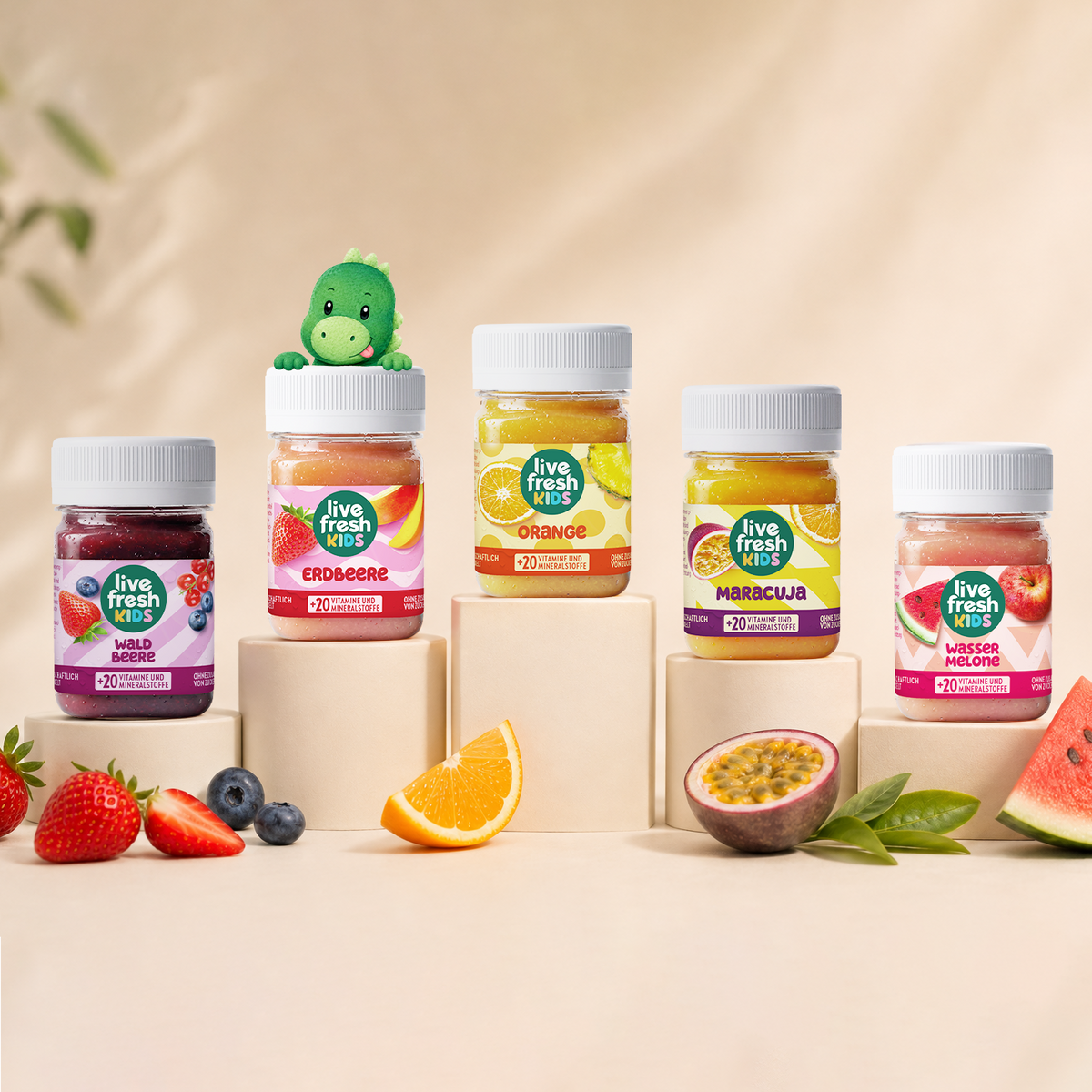 Five jars labeled “Live Fresh Kids,” “Wild Berries,” “Strawberry,” “Orange,” and “Passion Fruit.” "Watermelon" and "+20 Vitamins and Minerals" are displayed on pedestals of varying heights. A small green dinosaur peeks out from behind the strawberry jar, and fresh fruit lies in front of it.