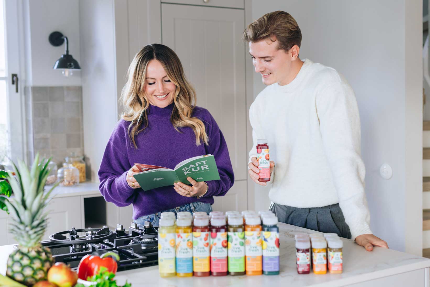 Eine junge Frau in einem lila Pullover und ein junger Mann in einem weißen Pullover stehen gemeinsam in einer modernen Küche. Die Frau hält ein grünes Buch mit dem Titel „SAFT KUR“ und lächelt, während der Mann interessiert zusieht und eine rote Flasche Live Fresh Saft in der Hand hält. Auf der Kücheninsel vor ihnen stehen mehrere bunte Saftflaschen in einer Reihe, umgeben von frischem Obst und Gemüse wie Ananas, Paprika und Äpfeln.