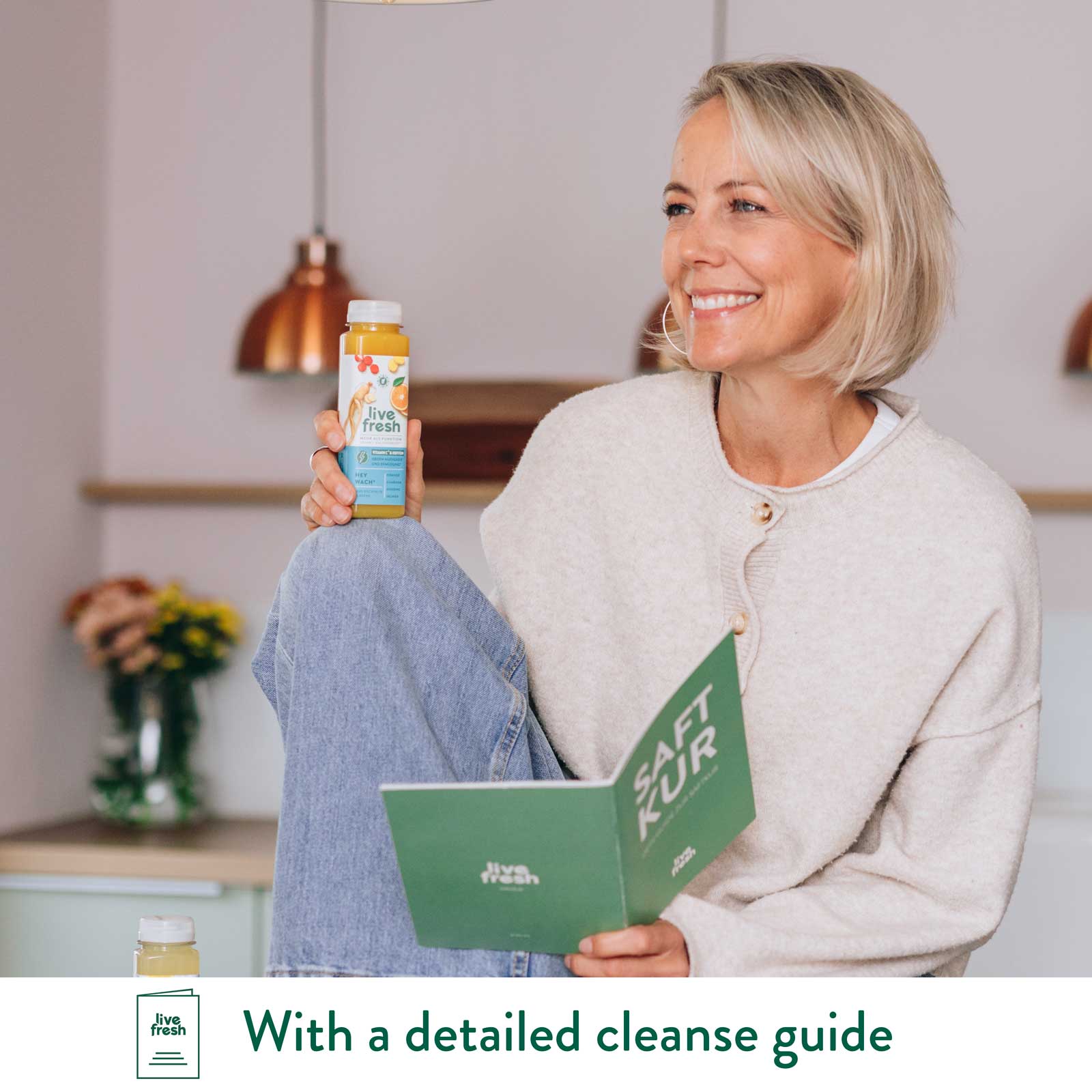 Smiling woman holding LiveFresh juice bottle while reading a cleanse guide in a cozy kitchen setting.