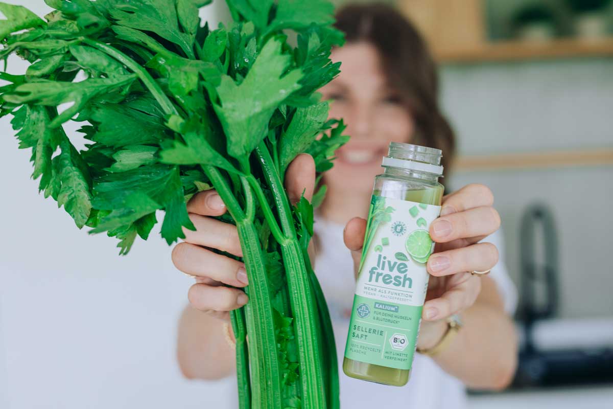 Frau hält frischen Staudensellerie in einer Hand und eine Flasche LiveFresh Selleriesaft mit Limette in der anderen Hand, ihr Gesicht ist verschwommen im Hintergrund zu erkennen. 