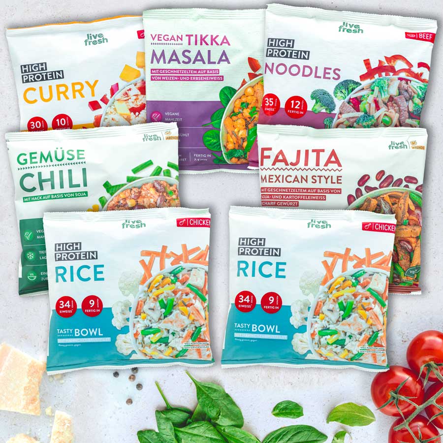 Flatlay mit sieben LiveFresh Tiefkühlgerichten, darunter High Protein Curry, Vegan Tikka Masala, Gemüse Chili, Fajita Mexican Style, High Protein Noodles und zwei High Protein Rice Varianten. Umgeben von frischem Spinat, Tomaten und Parmesanstücken.