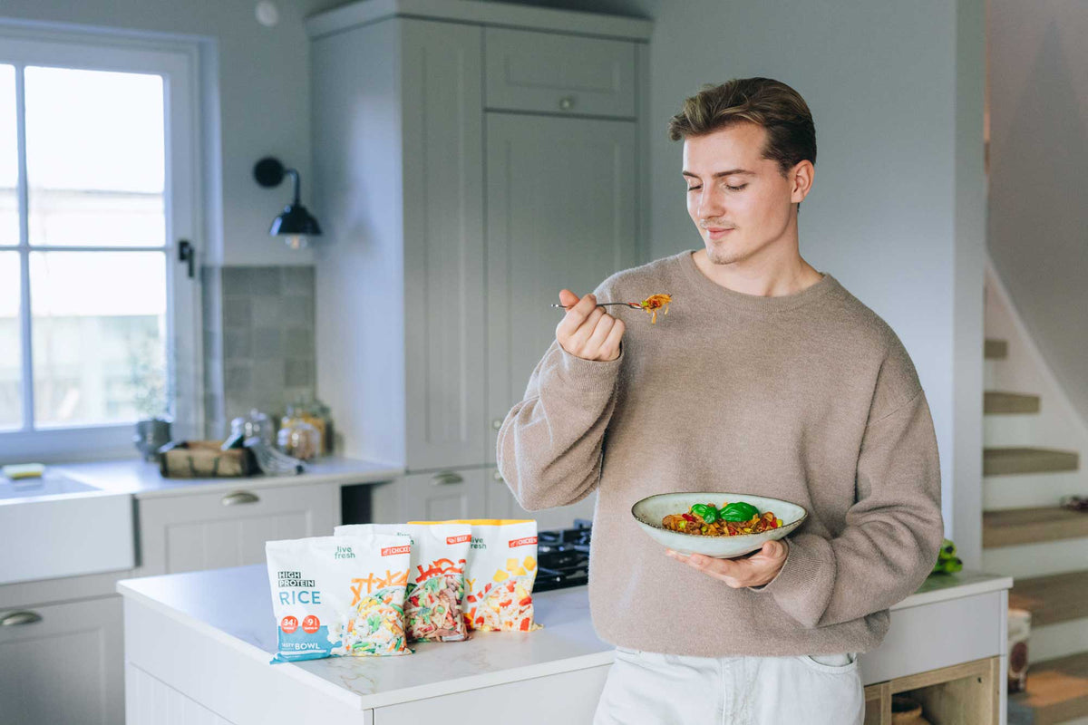 Junger Mann in heller Küche probiert ein Fertiggericht aus einer Schüssel, vor ihm stehen weitere  Fertiggerichte von LiveFresh. 