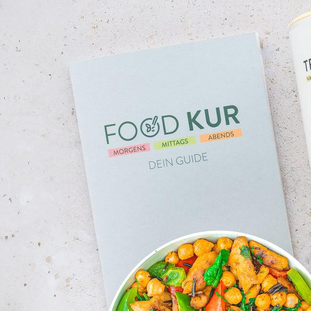 Heft mit der Aufschrift „Food Kur – Morgens, Mittags, Abends – Dein Guide“ auf einer hellen Oberfläche. Neben dem Heft ist eine Schüssel mit einem bunten Gericht zu sehen, das Gemüse wie Erbsenschoten, Kichererbsen und Paprika enthält.