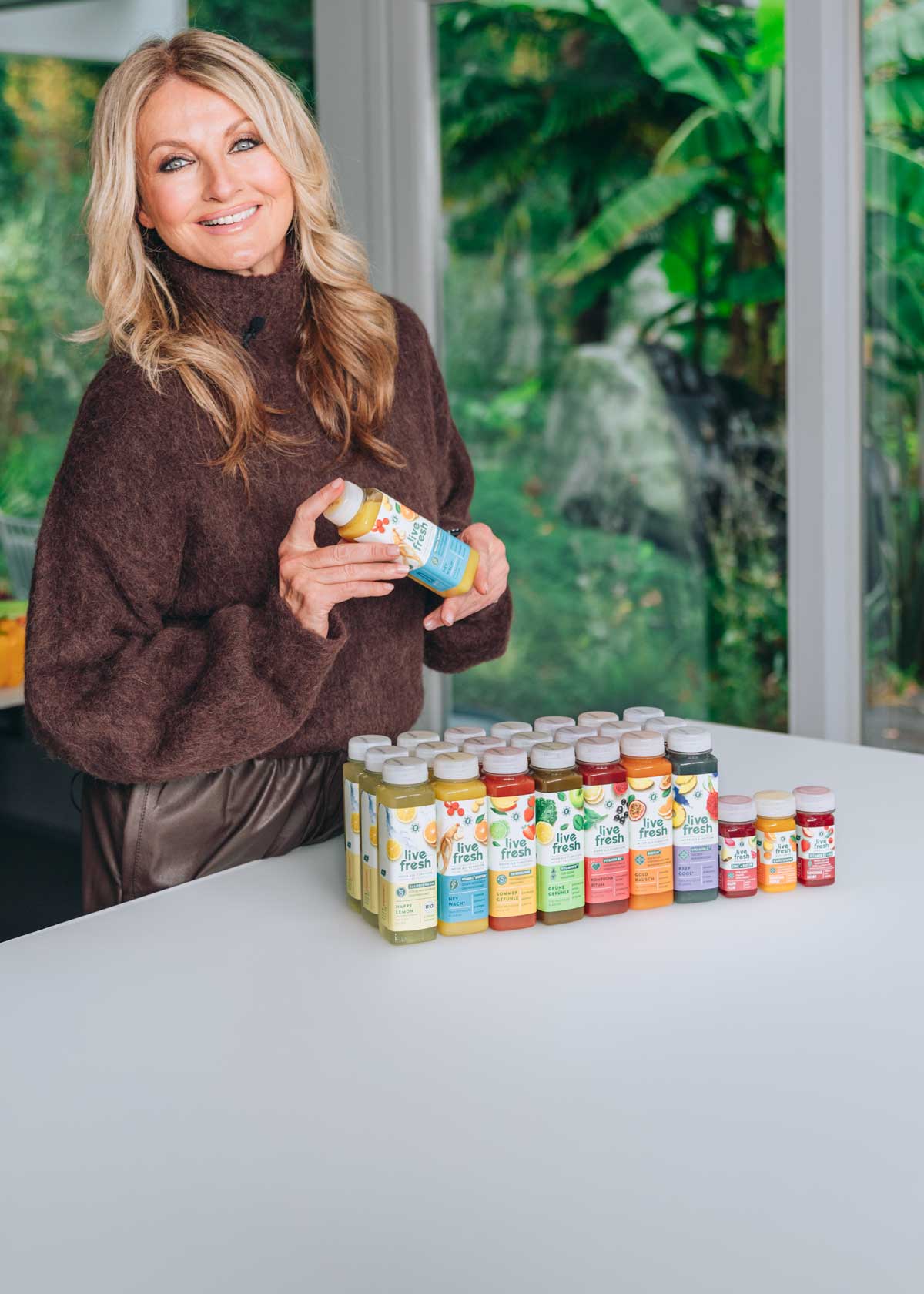 Blonde Frau (Frauke Ludowig) in braunem Pullover steht in moderner Küche und präsentiert mehrere bunte Saftflaschen von der kaltgepressten LiveFresh Saftkur auf einer weißen Kücheninsel.