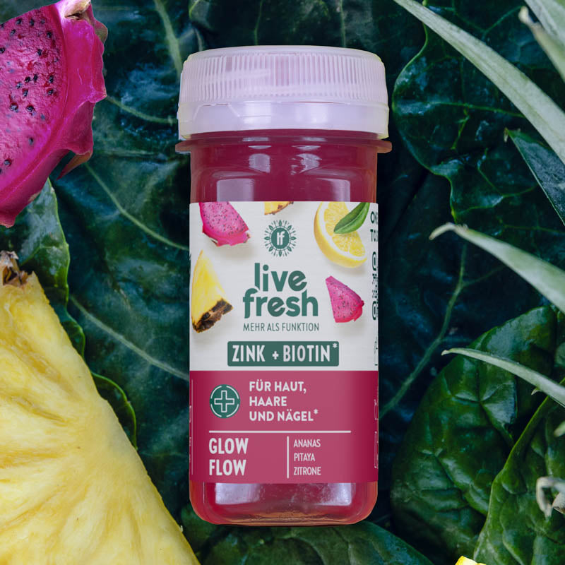 LiveFresh Shot „Glow Flow“ mit Zink und Biotin, beworben für Haut, Haare und Nägel. Umgeben von Ananas, Pitaya und Zitrone, mit sattem pink-rotem Farbton vor grünem Blattgemüse.