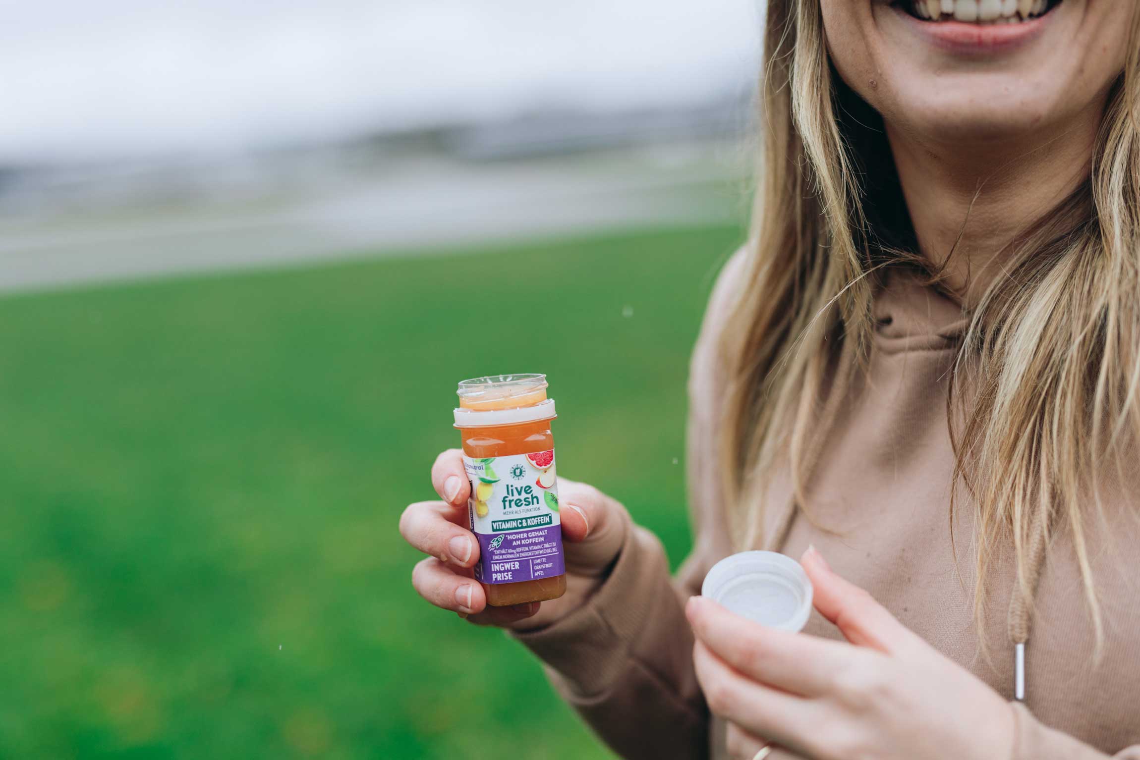 Person hält lächelnd einen geöffneten Shot namens "Ingwer Prise" von LiveFresh  in der Hand, vor grünem Hintergrund im Freien.