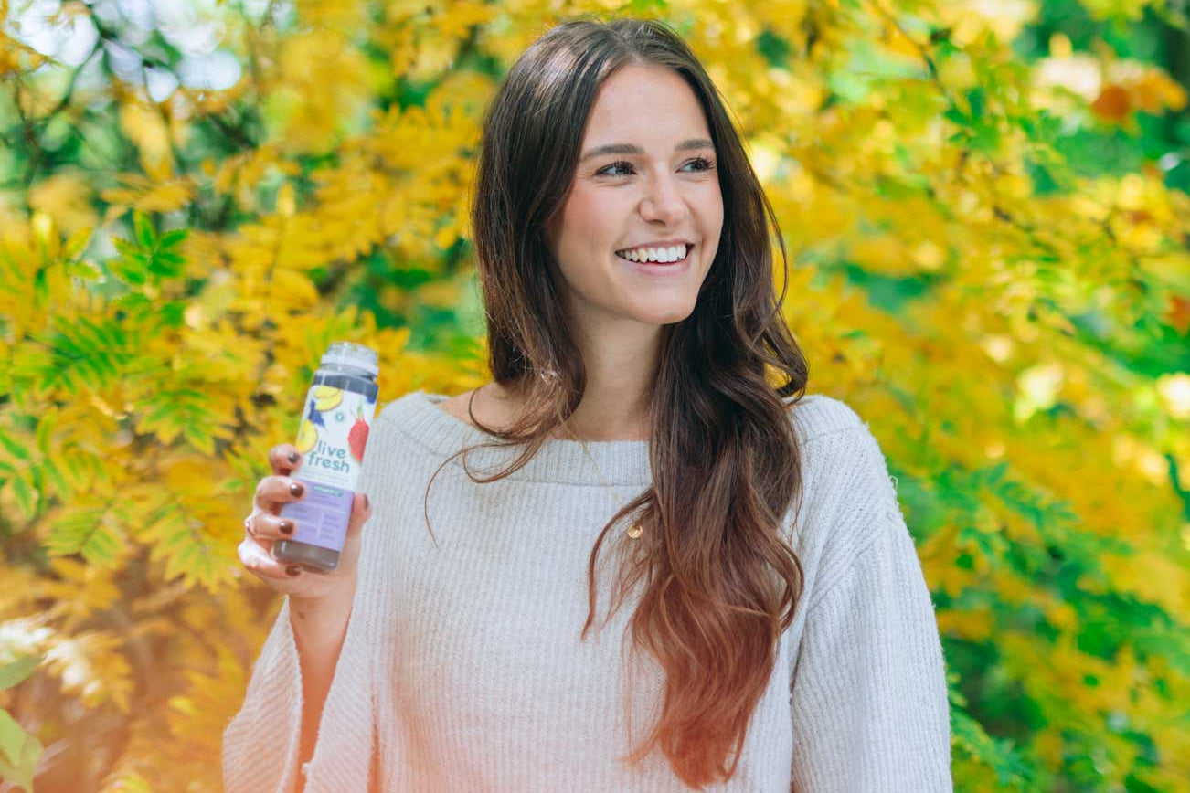 Lächelnde Frau mit langen braunen Haaren hält eine Flasche kaltgepressten Saft namens Keep Cool vor leuchtend gelbem Herbstlaub.