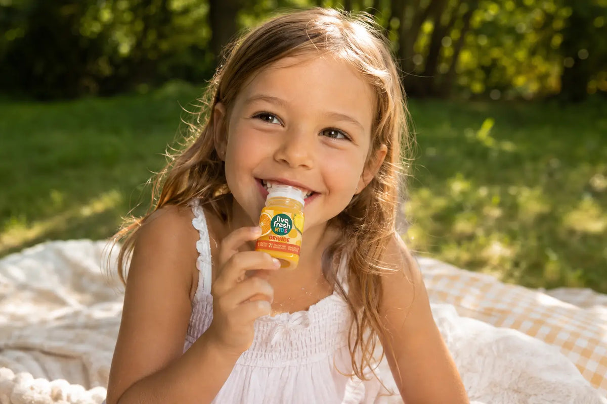 A girl sitting on a picnic blanket in a green space is smiling as she drinks a LiveFresh Kids orange juice. 