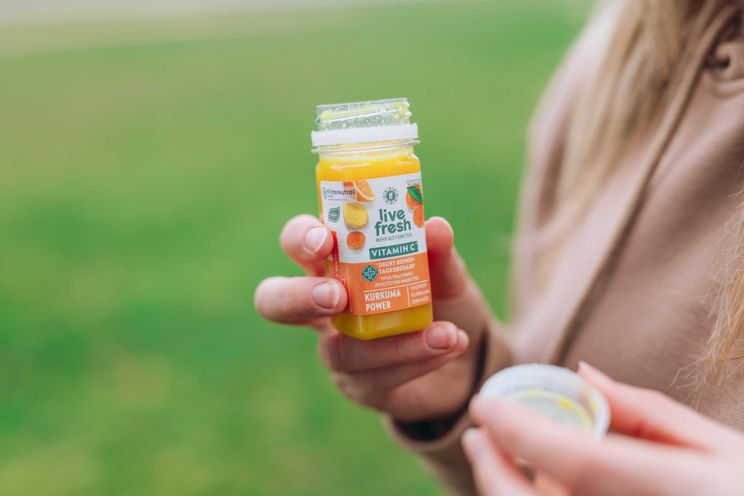 Eine Person, die einen Kurkuma Power Shot von Live Fresh in der Hand hält. Der Shot verspricht Vitamin C und enthält Zutaten wie Ingwer, Kurkuma und Orange. Die Person hält den geöffneten Deckel in der anderen Hand, während sie den Shot draußen, vor einem grünen Hintergrund, genießt.