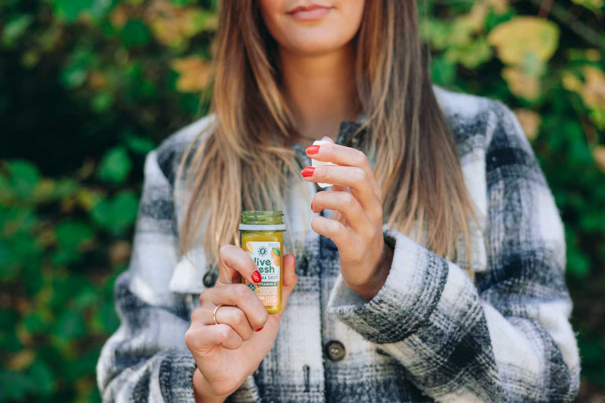 eine Frau, die draußen steht und eine Flasche mit einem Kurkuma-Shot von LiveFresh in der Hand hält. Sie öffnet den Deckel der kleinen Flasche, während ihre roten Nägel im Kontrast zu ihrem karierten Mantel und dem natürlichen grünen Hintergrund auffallen. 