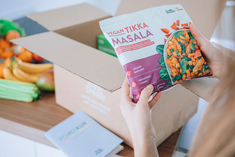 Tiefkühlgericht „Vegan Tikka Masala“ von LiveFresh wird von einer Person aus einem Versandkarton entnommen. Im Hintergrund liegen frische Lebensmittel auf einer Küchenarbeitsplatte. Verschwommen ist ein grauer Foodkur-Guide neben dem Karton zu erkennen. 